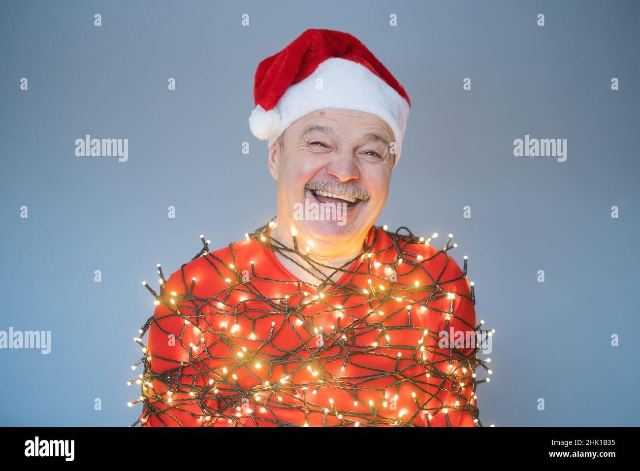Uomo anziano in rosso cappello di natale avvolto in ghirlanda ridendo essere felice. Nuovo anno concetto 2023 Foto Stock