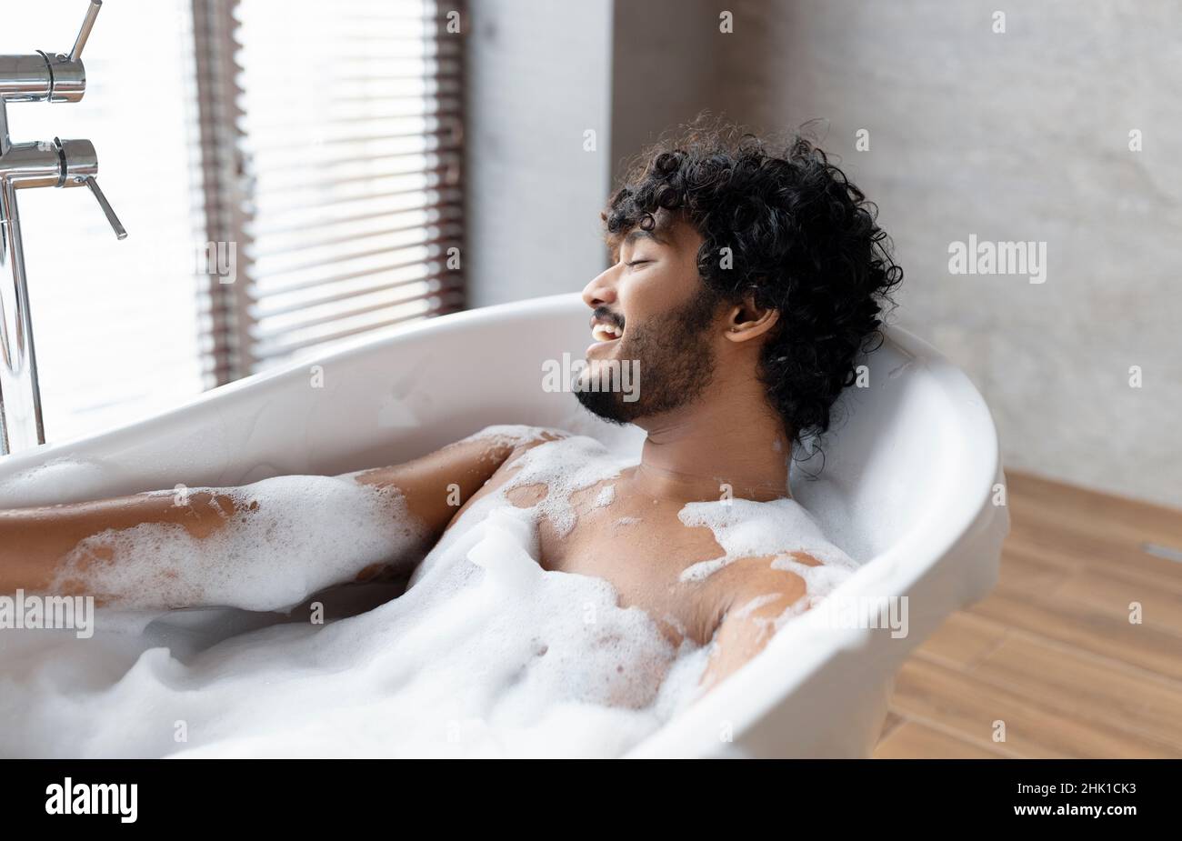Bagno rilassante. Uomo indiano sdraiato nella vasca da bagno, godendo di schiuma a casa spa, riposo in acqua calda a casa, spazio libero Foto Stock
