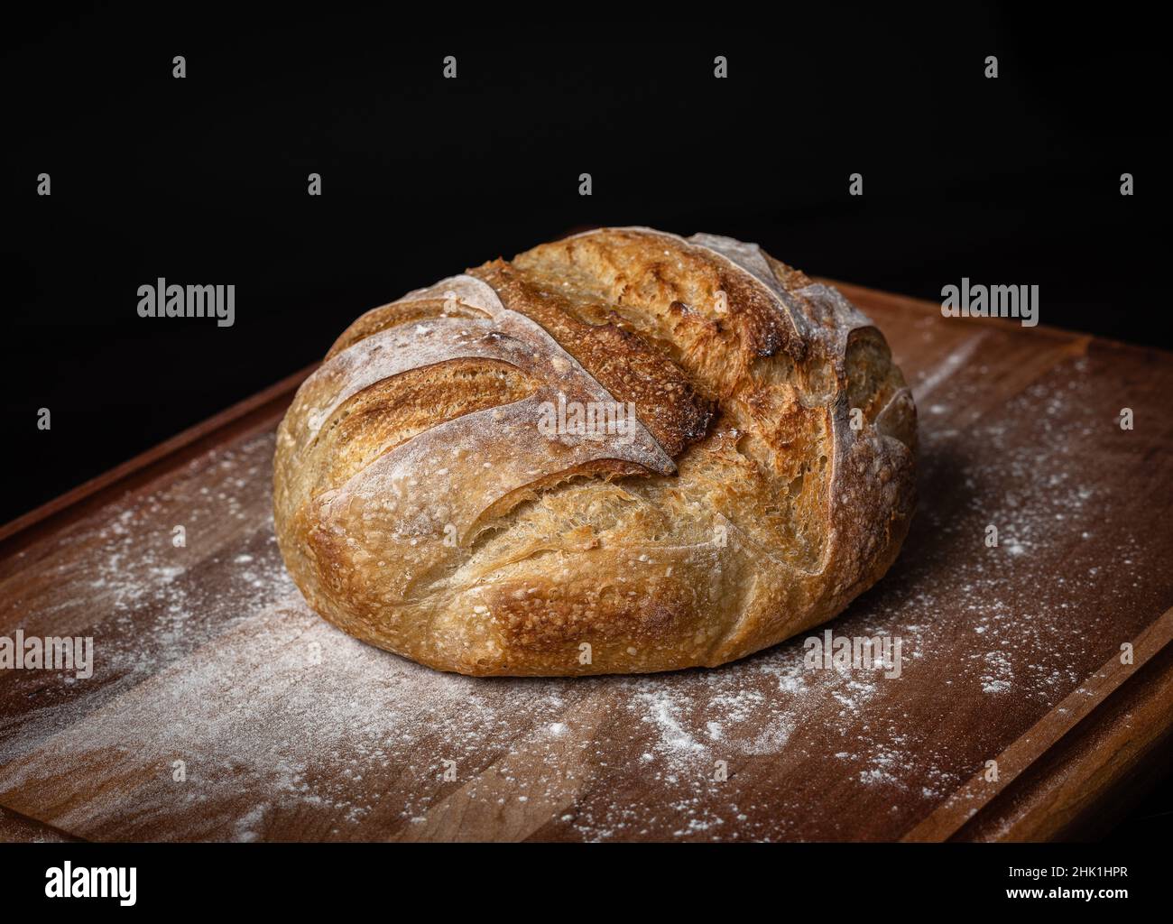 Una pagnotta di pane di pasta madre appena sfornata in farina su una tavola con fondo nero. Foto Stock