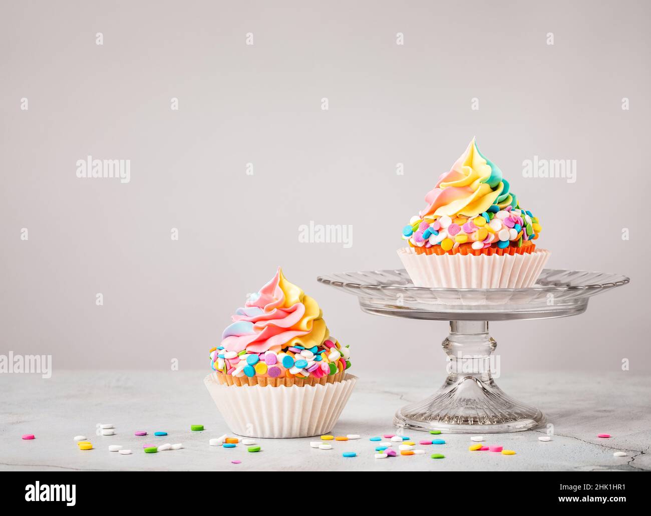 Due cupcake Rainbow Birthday con colorate spolverate su sfondo grigio chiaro. Foto Stock