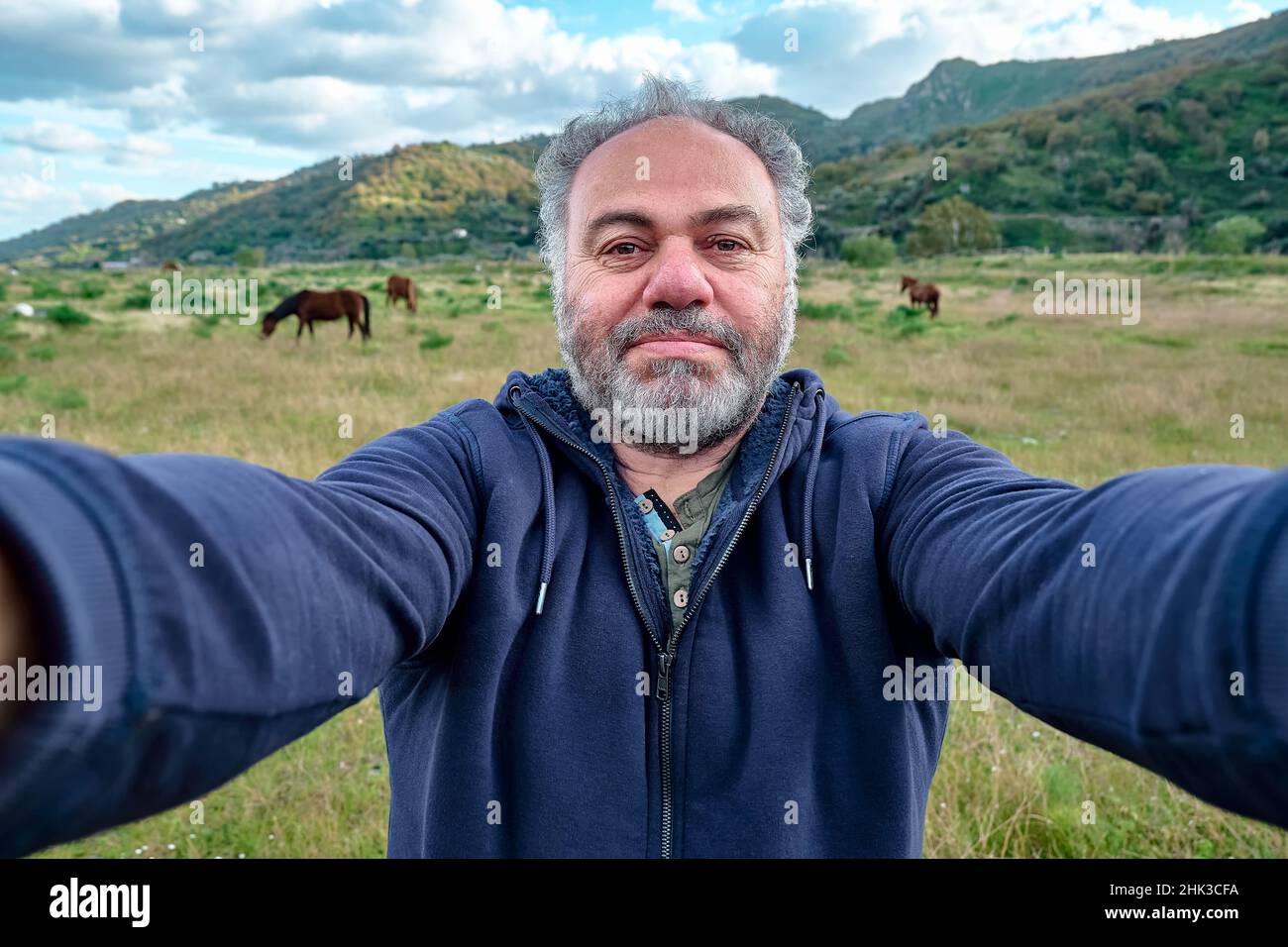 Felice, serio uomo maturo bearded che fa un selfie sul prato con i cavalli di pascolo. Concetto di attività ricreative, turismo, stile di vita e natura. Foto Stock