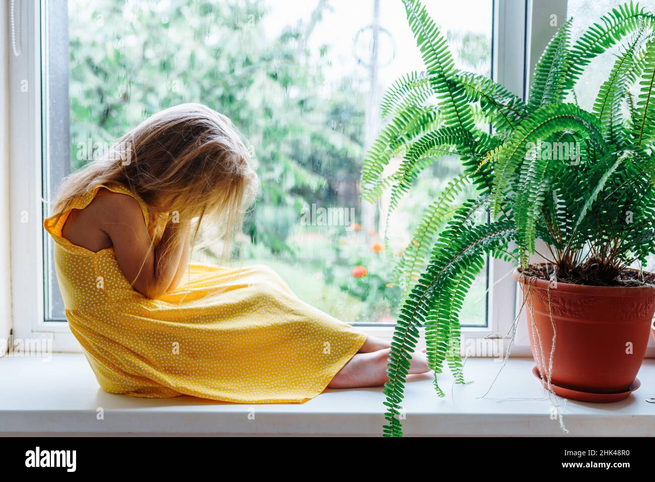 Giovane ragazza piangendo a piedi nudi seduta sul davanzale della finestra in mani vicino pianta felce in vaso di argilla. Vista laterale. Foto Stock