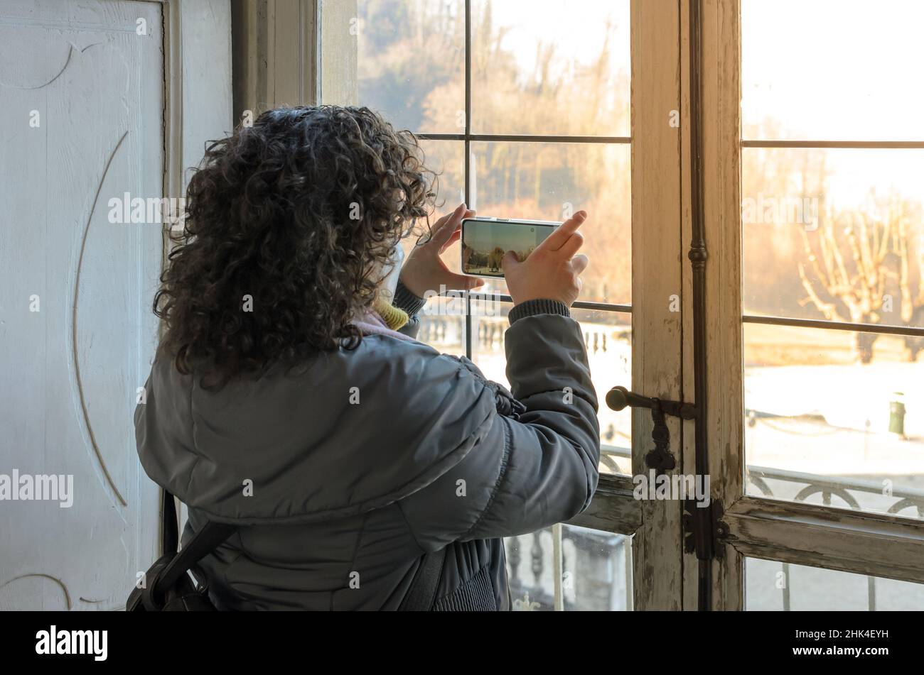 Donna turistica che fa una foto del paesaggio esterno con il suo telefono attraverso il vetro di una vecchia finestra di legno di una casa antica. Foto Stock