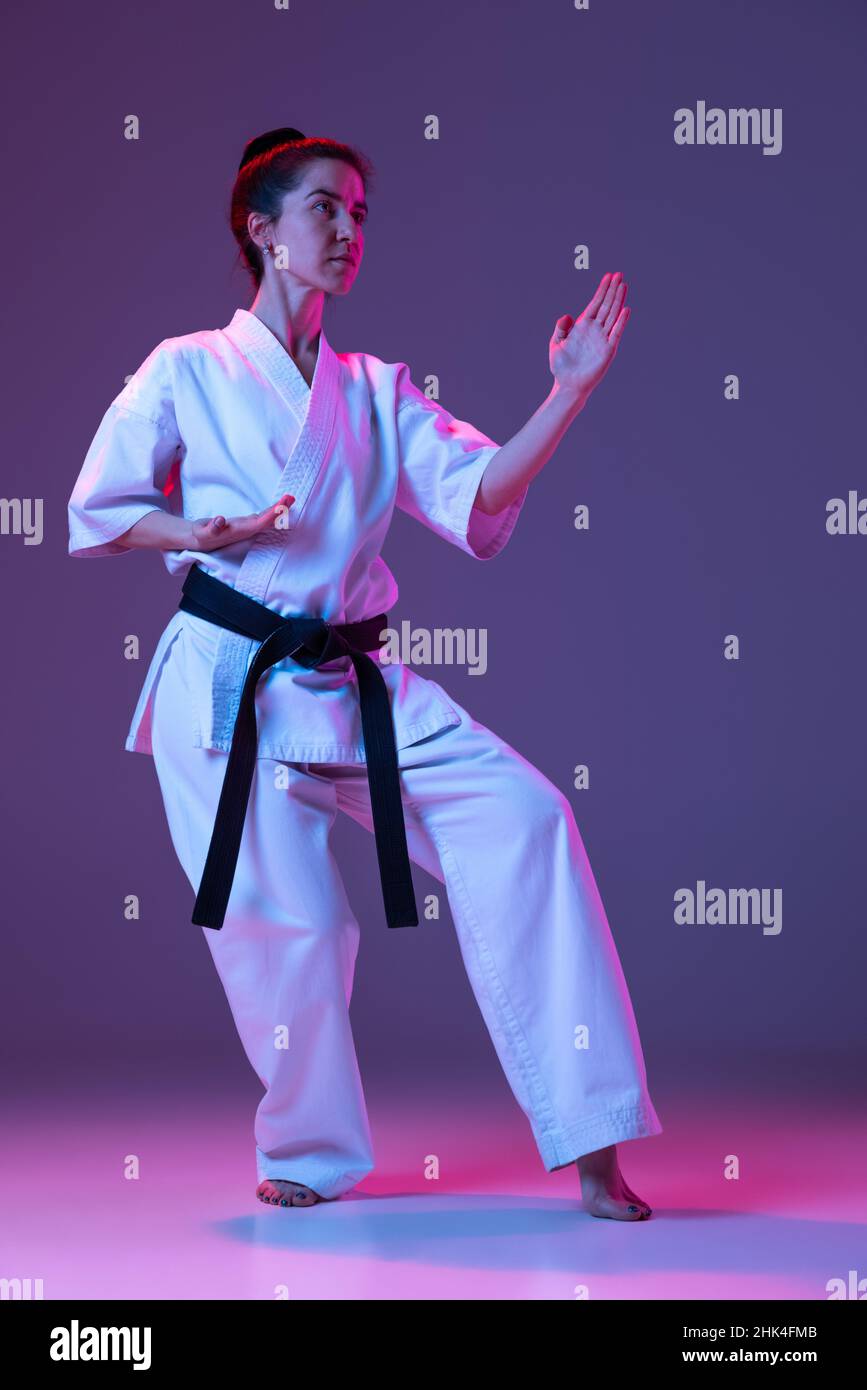Ritratto di taekwondo femminile, karate atleti in doboks che fanno movimenti di base isolato su sfondo viola in neon. Concetto di sport, arti marziali Foto Stock