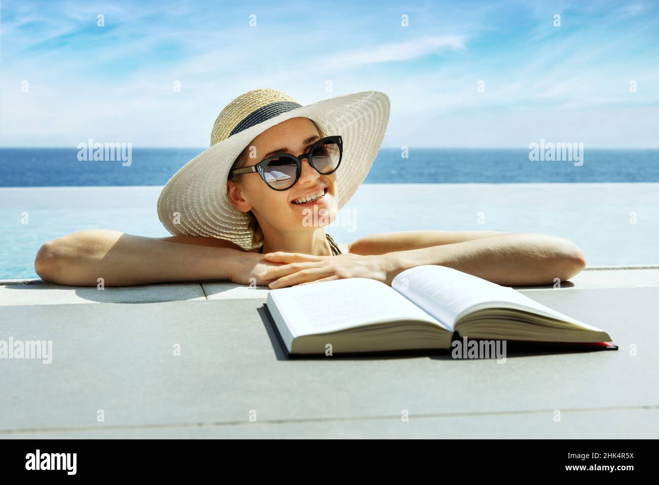 donna sorridente e attraente con cappello e occhiali da sole leggendo un libro e rilassandosi nella piscina a sfioro presso il resort di vacanza Foto Stock