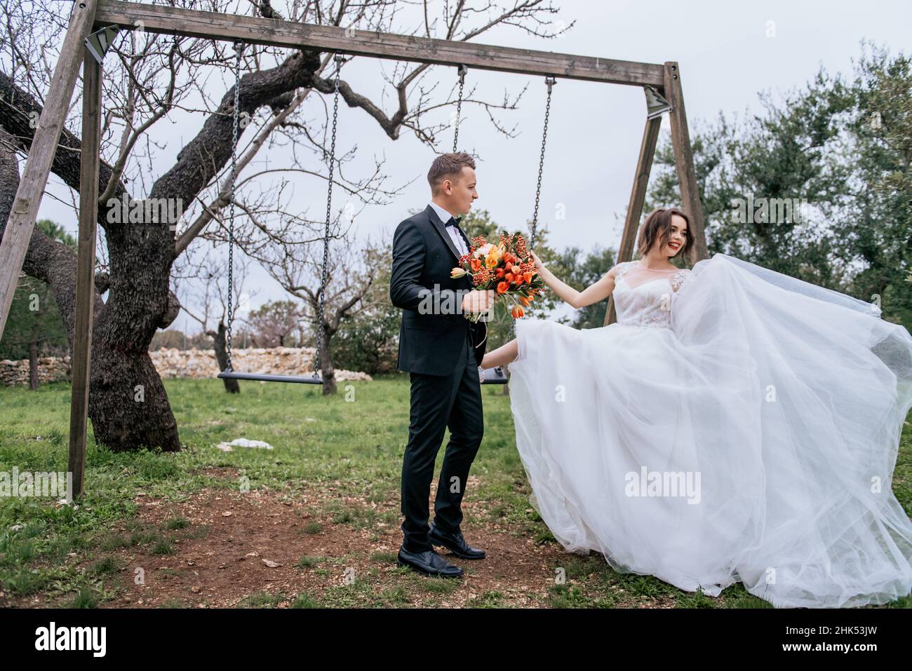 Felice coppia sorridente di stile che cammina in Toscana, Italia il giorno del matrimonio. SPOSI NOVELLI CON L'ALTALENA NEL PARCO. La sposa e lo sposo camminano giù il Foto Stock