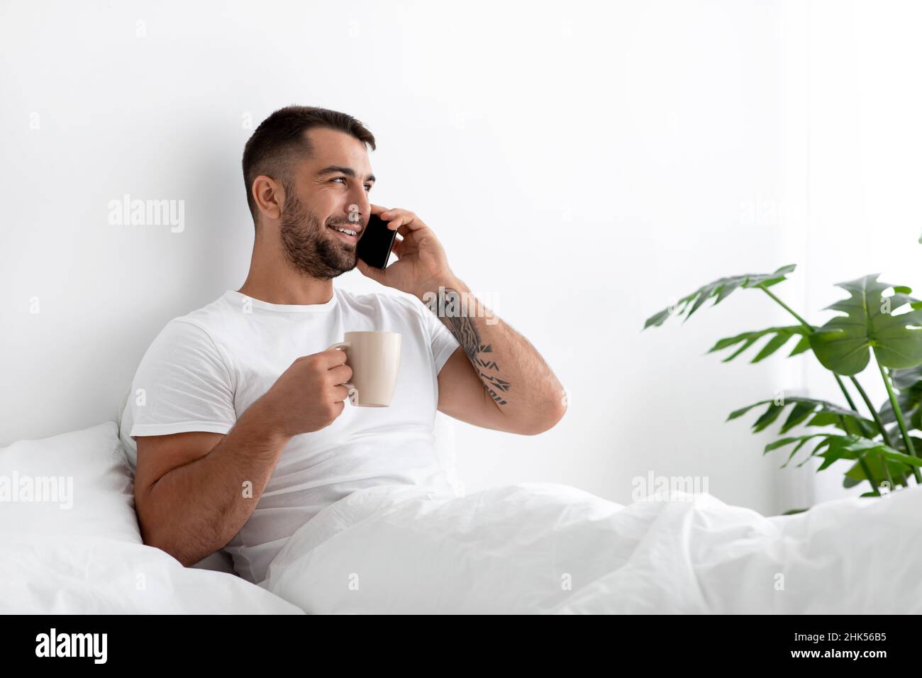 Sorridente millennial muscolare europeo beve caffè, chiama da smartphone e gode di buona mattina in camera da letto Foto Stock