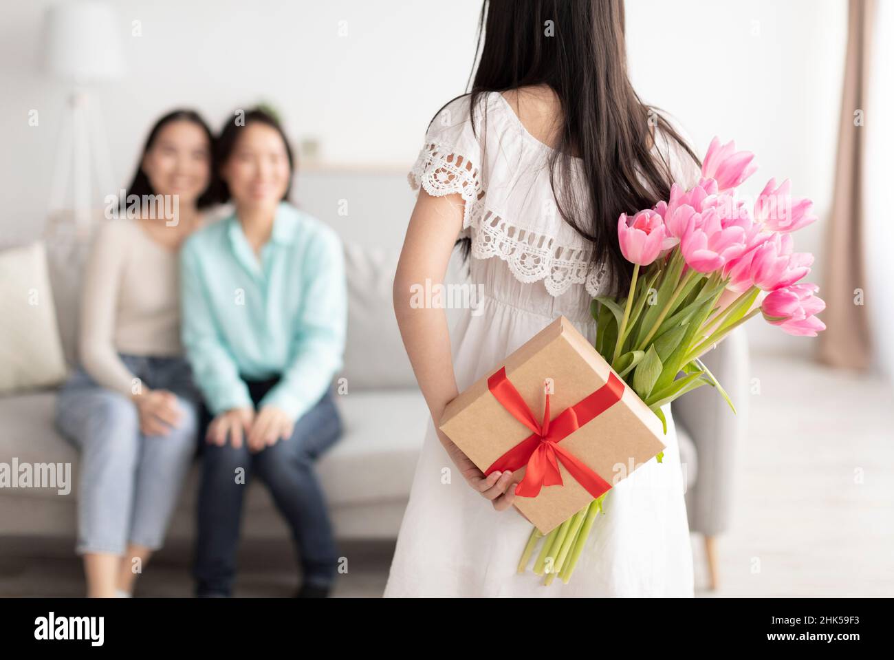 Festa della donna. Ragazza asiatica nascondendo dono e fiori dietro la schiena, congratulandosi con madre e nonna con vacanza Foto Stock