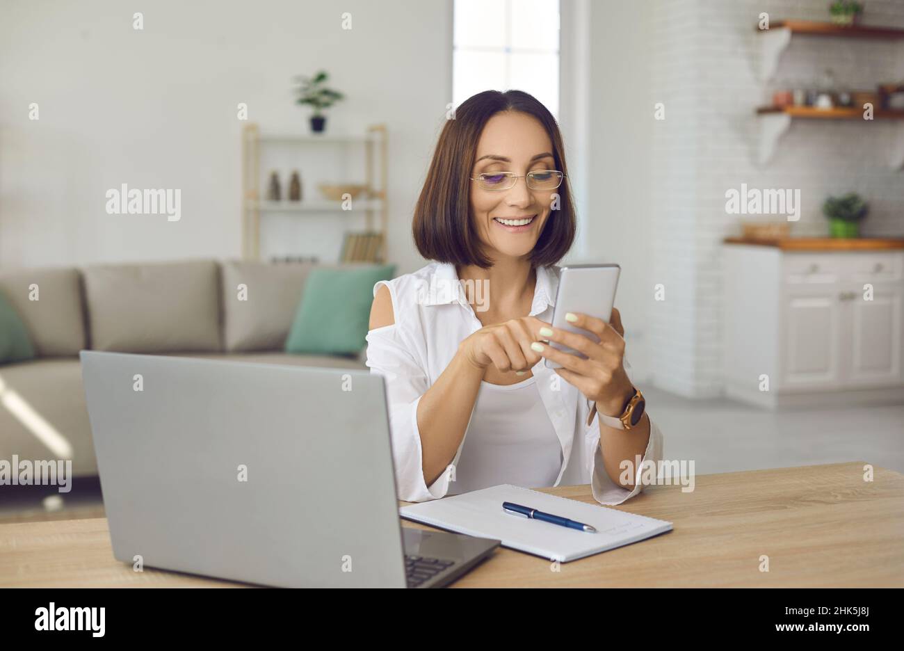 Donna felice che usa il telefono cellulare mentre si siede alla scrivania con il portatile e il notebook a casa Foto Stock