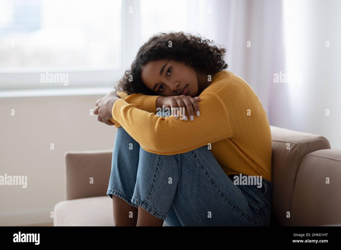 Giovane donna nera depressa che abbraccia le ginocchia sul divano a casa, assorbita in pensieri negativi, avendo disturbi mentali Foto Stock