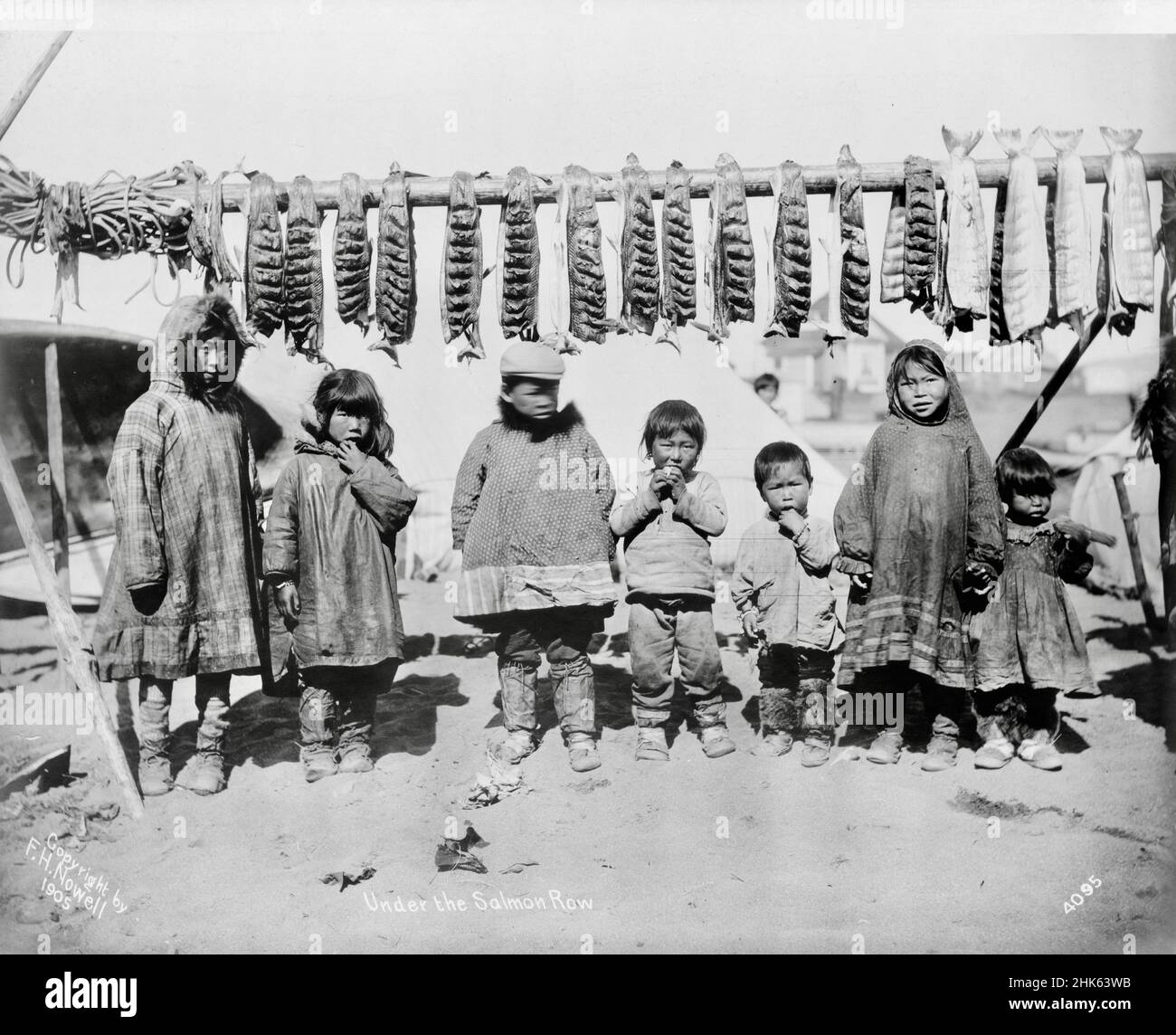 Frank H Nowell fotografia dal titolo The Salmon Row - salmone appeso da una rastrelliera sopra una fila di bambini Inuit. Foto Stock