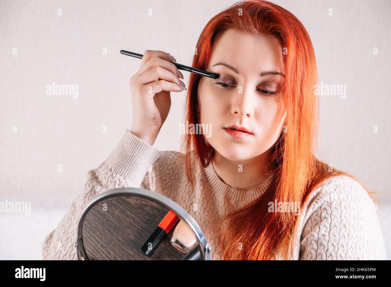 Giovane donna con capelli rossi applica trucco. La signora guarda nello specchio e usa i cosmetici. Foto Stock