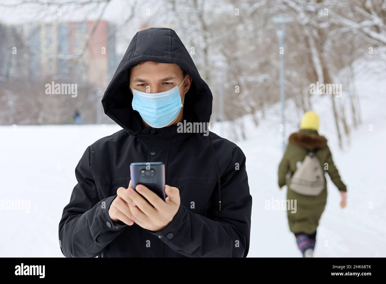 Protezione da coronavirus, uomo in maschera medica su una strada invernale della città con smartphone in mano. Concetto di sicurezza durante il covid-19 pandemia, tempo di neve Foto Stock