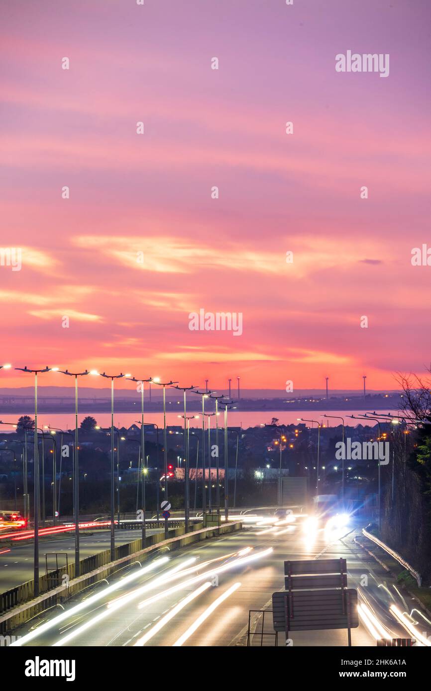 Spettacolare cielo rosso serale dopo il tramonto sulla Thanet Way, A299, una strada a due corsie che collega il Nord Est del Kent con il M2 e Londra. Sullo sfondo si trova l'isola di Sheppey, alcune turbine eoliche e l'estuario del Tamigi. Ora di punta con traffico sulla strada. Sfocatura del movimento con i percorsi dei fari. Foto Stock