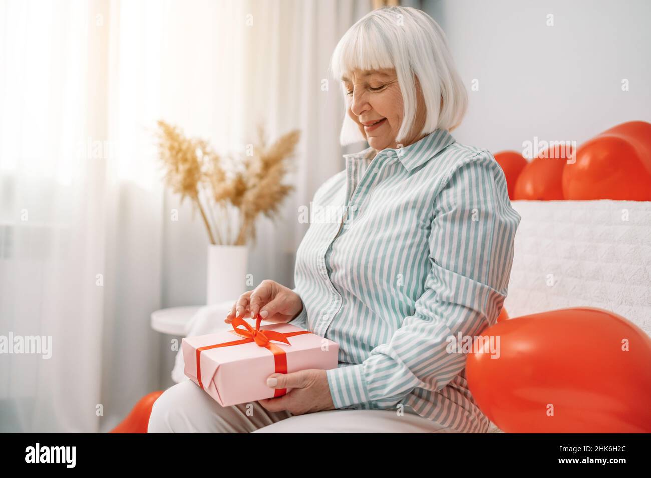San Valentino, giorno della mamma. Donna anziana con capelli biondi di 40s 50s anni che indossa elegante blusa aperta su carta rosa regalo sorpresa presente con ribo rosso Foto Stock