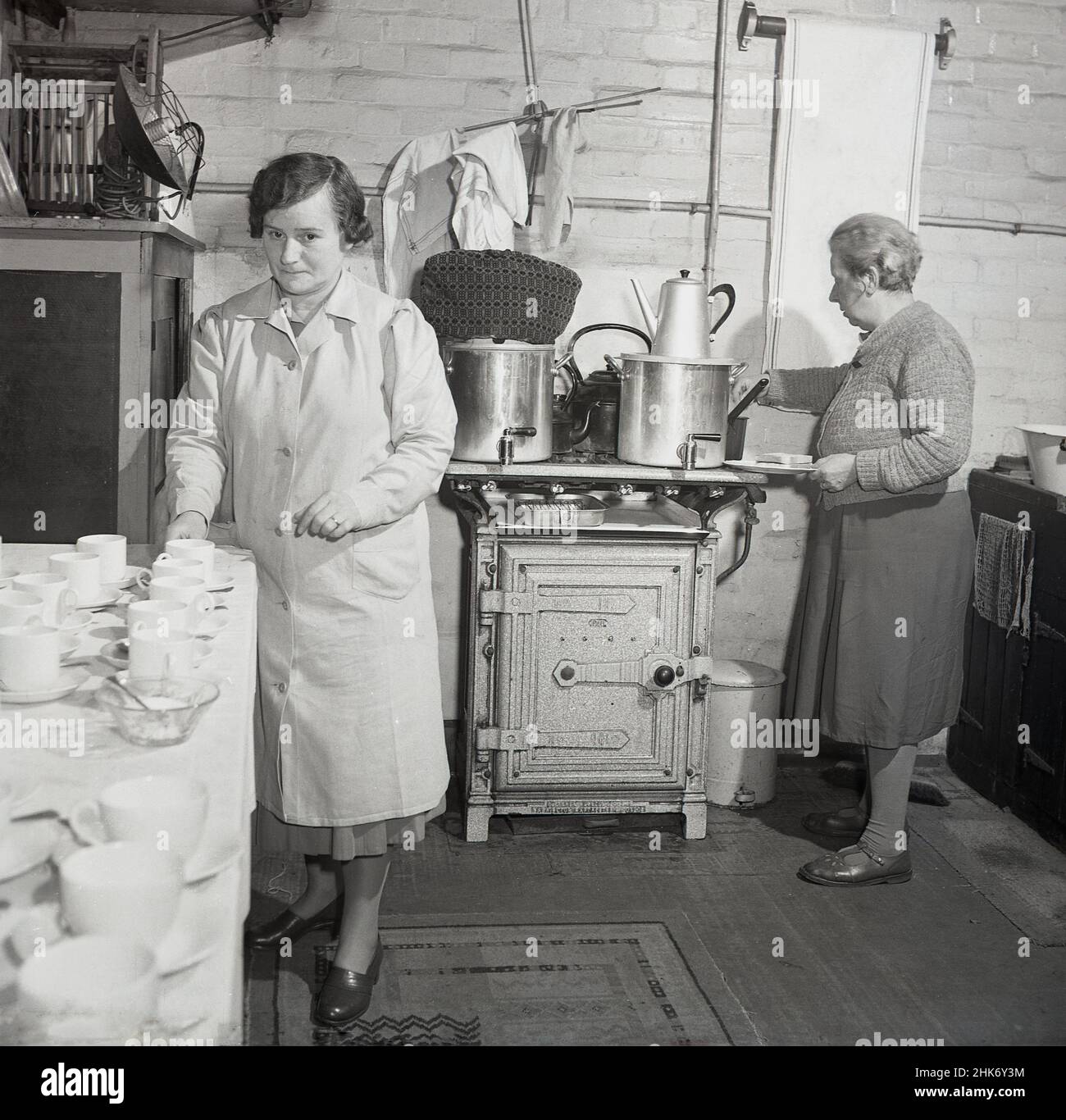 1950s, Inghilterra. Due Signore che fanno il tè in una cucina di fabbrica, in questo quadro storico da J Allan Cash, Inghilterra, Regno Unito. Sulla vecchia stufa è due urne del tè ed uno del lavoratore di ristorazione anziano sta tenendo un piatto con una fetta di pane sopra. Foto Stock