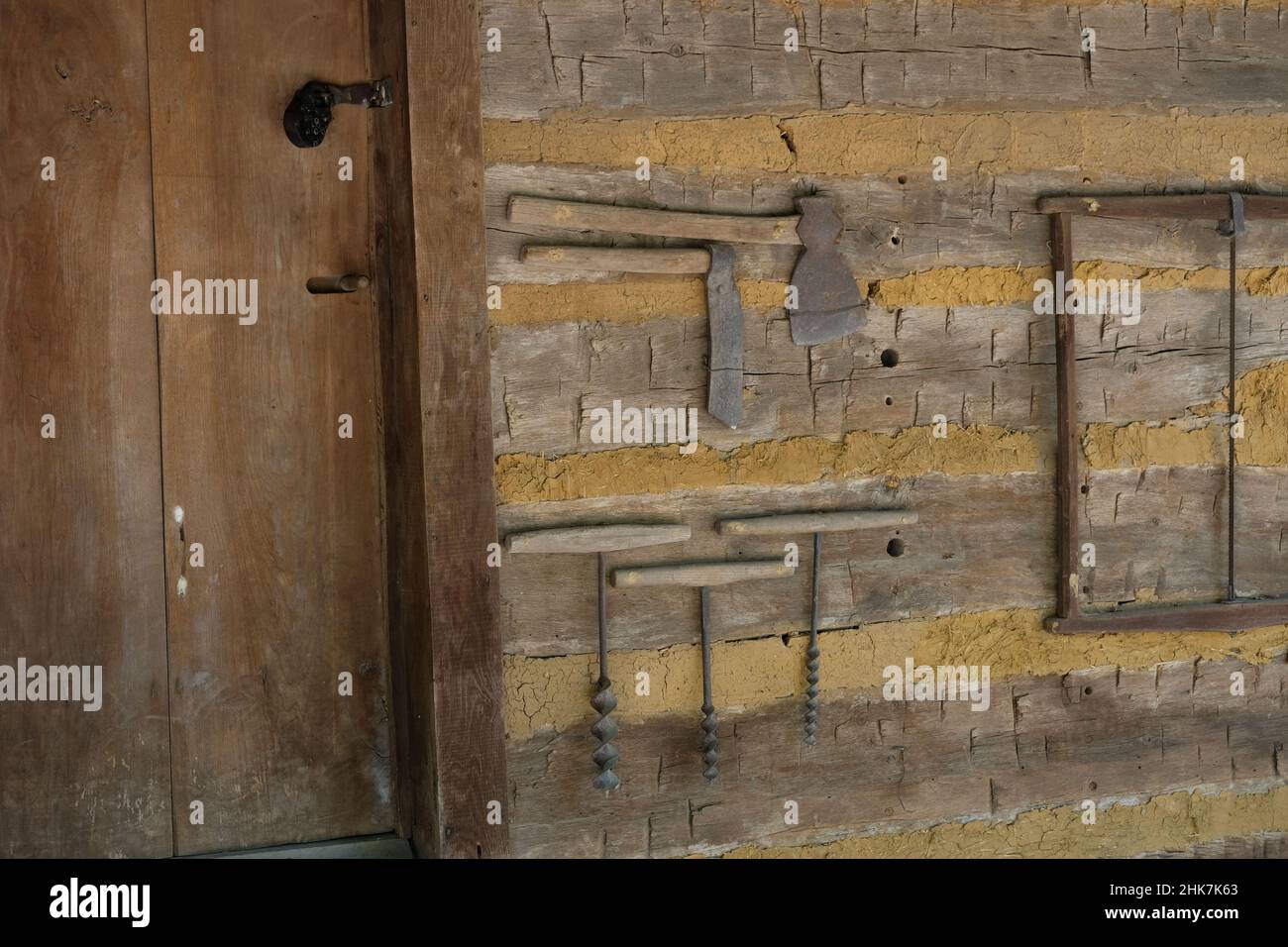 Vecchi attrezzi legno e ferro, a mano, assi corti e lunghi, trapani a mano di diametro variabile. Appeso su legno e muro di terra accanto alla porta di legno chiusa. Foto Stock