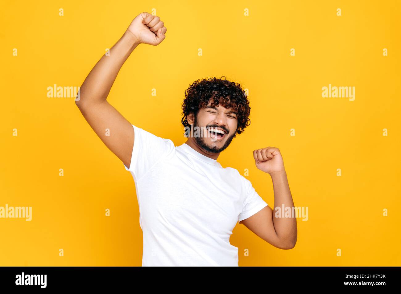Estatica fortunato felice ricurly indiano uomo, ballando, gesturing con pugni, riceve profitto, felice di vincere la lotteria, in piedi su sfondo arancione isolato. Buone notizie, successo e concetto di felicità Foto Stock