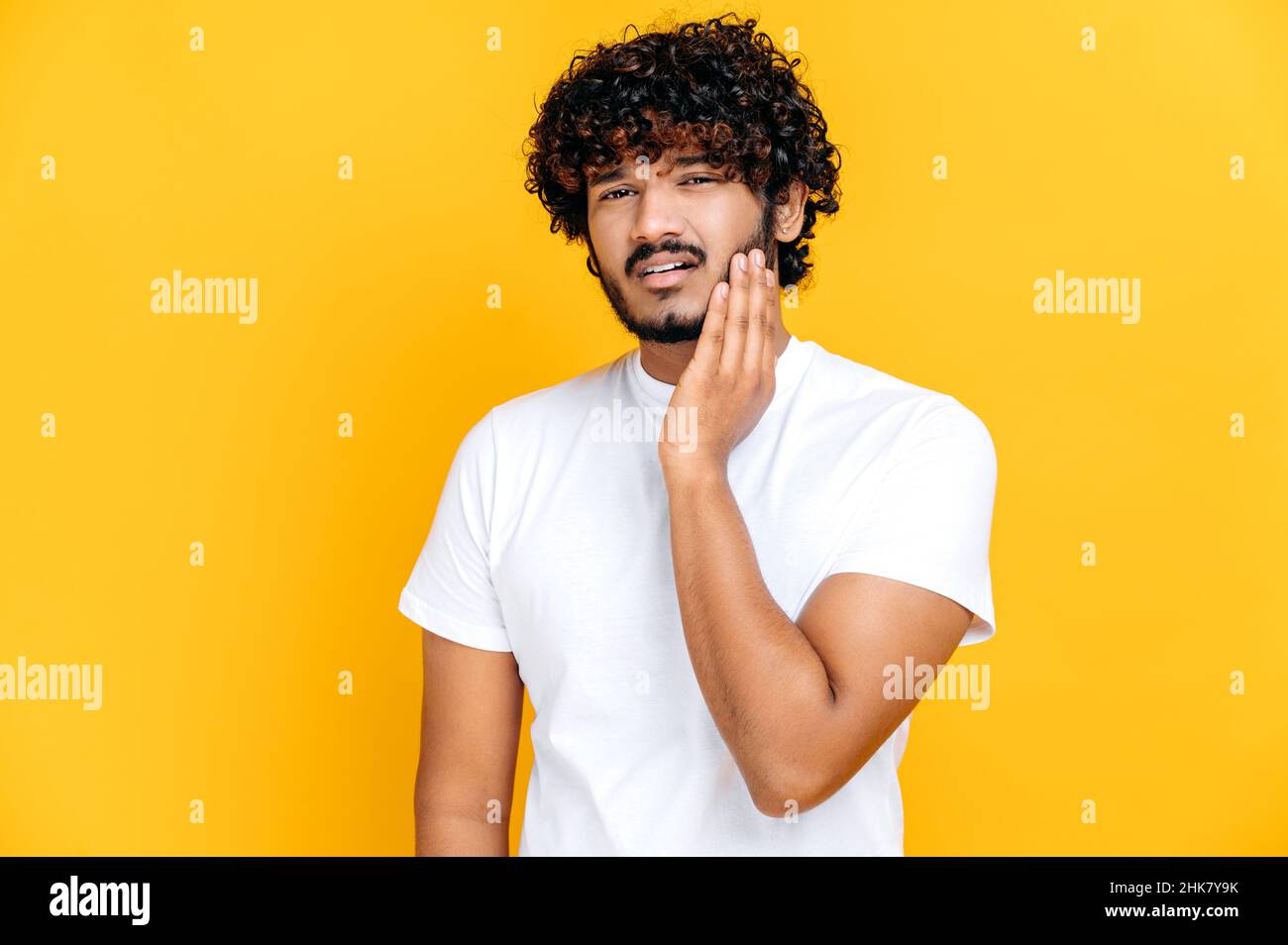 Concetto di mal di denti. Triste malsano indiano o arabo ragazzo toccare guancia con mano con espressione dolorosa a causa di mal di denti o malattie dentali sui denti, in piedi su sfondo arancione isolato Foto Stock