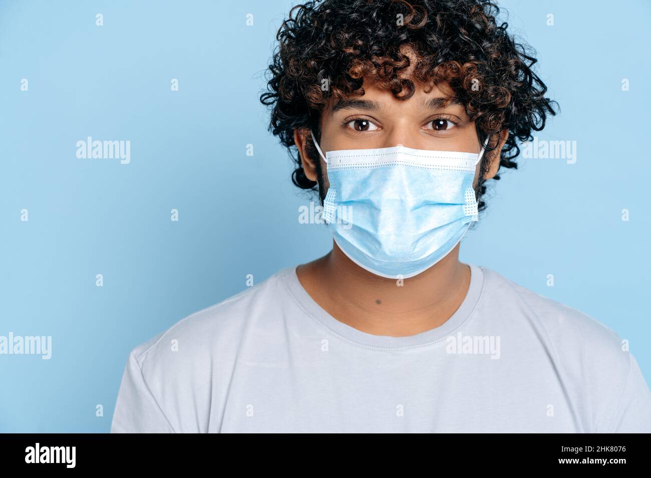 Prevenzione delle infezioni. Primo piano di un ragazzo curly indiano o arabo giovane adulto, con una maschera medica protettiva sul suo volto, guardando la fotocamera mentre si trova su un isolato sfondo blu Foto Stock