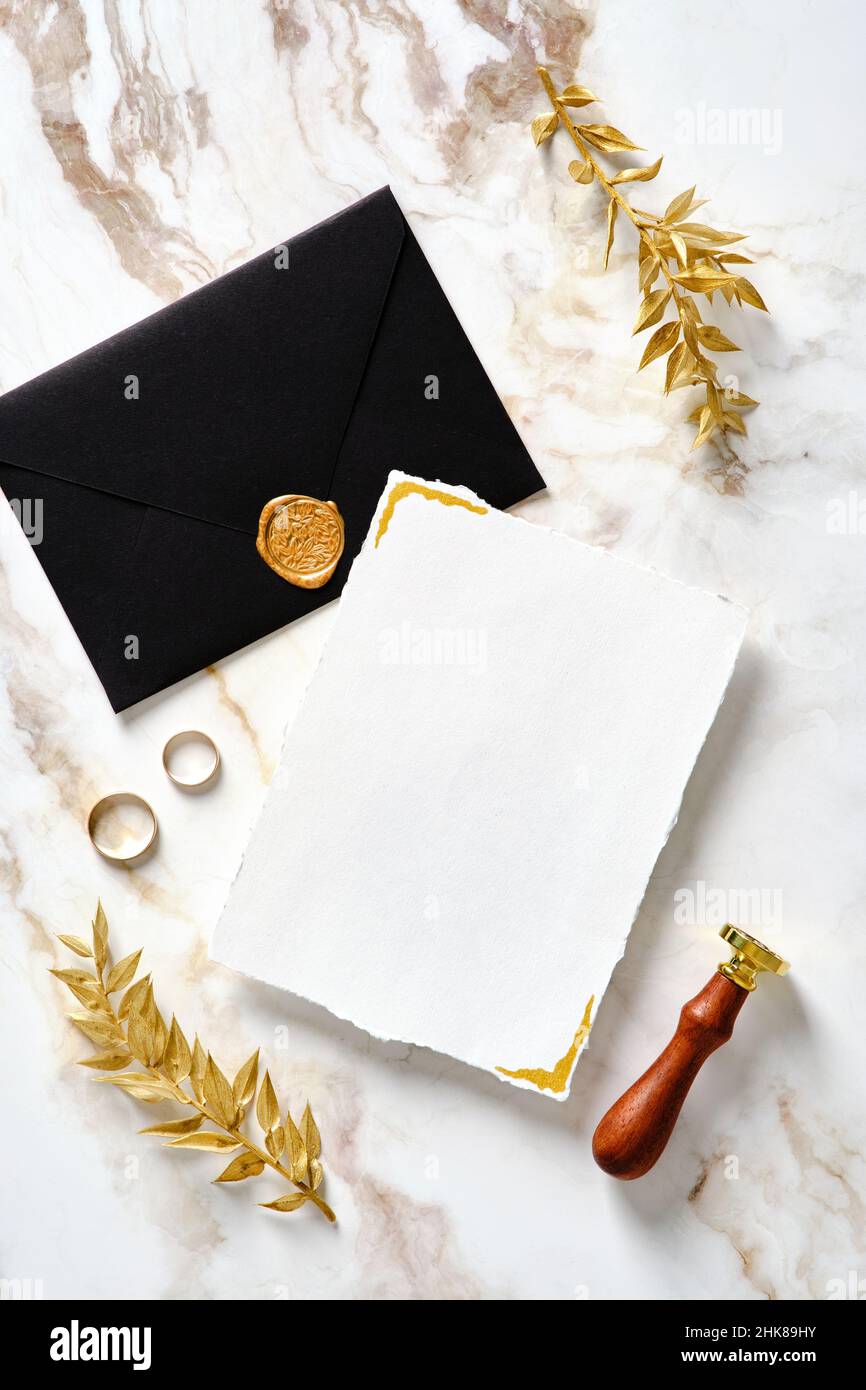 Mockup di cancelleria di nozze. Carta di invito per matrimoni vuota con busta nera, sigillo in cera, anelli dorati, foglie su sfondo oro marmo. Disposizione piatta Foto Stock
