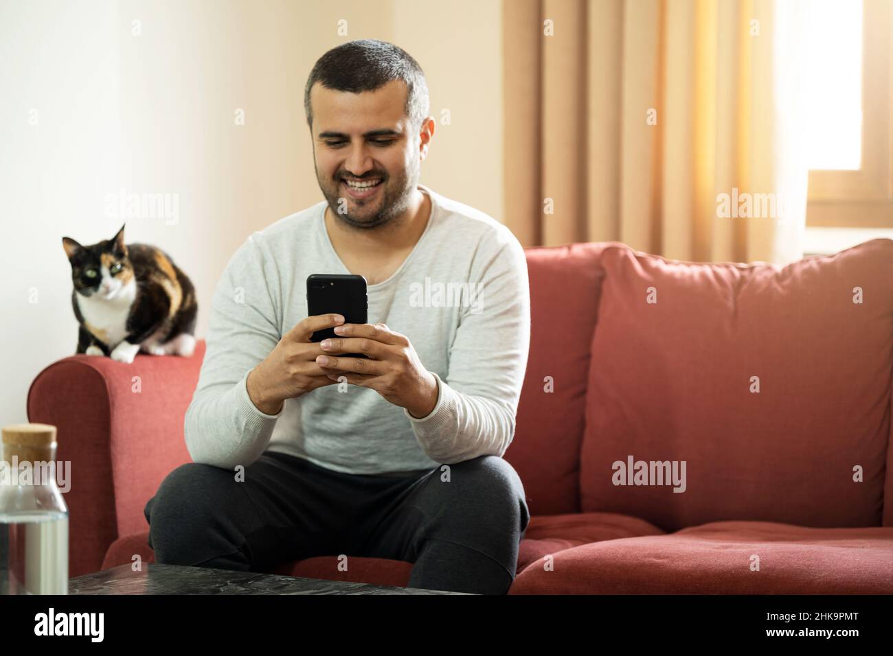 Bell'uomo dai capelli corti che tiene lo smartphone e si siede sul divano con il gatto. Spazio di copia Foto Stock