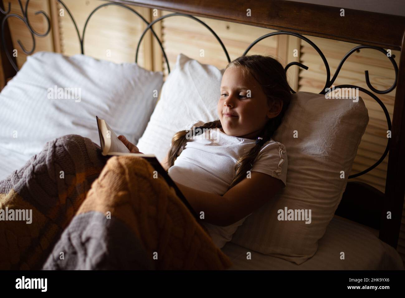 Curiosa bambina femminile sdraiata su un enorme letto matrimoniale con libro in mano che guarda il libro e legge indossando pigiami in casa di campagna in legno. Tempo libero Foto Stock