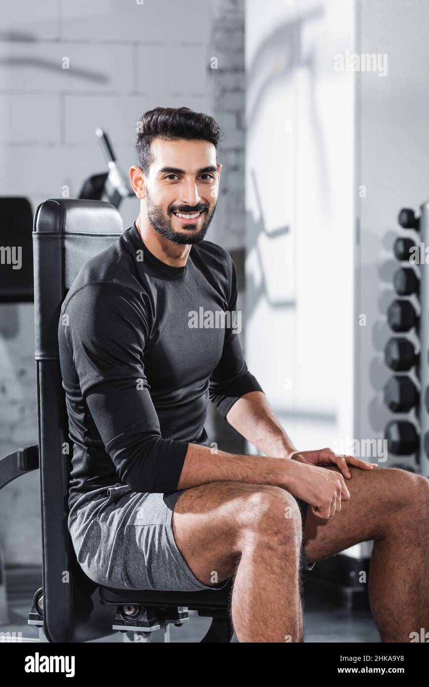 Sorridente sportivo arabo che guarda la macchina fotografica mentre si siede su attrezzature sportive in palestra Foto Stock