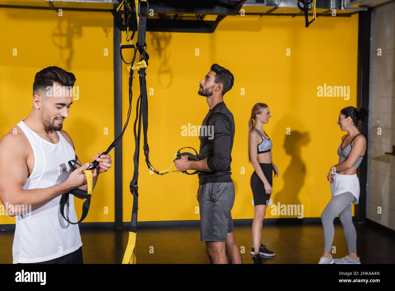 Sportivo sorridente che tiene le cinghie delle sospensioni vicino a amici multietnici sfocati in palestra Foto Stock