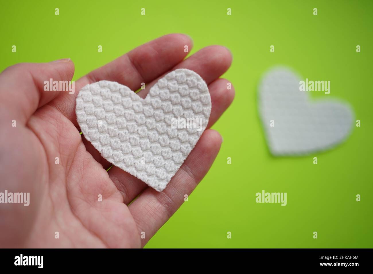 Mano con cuore in tessuto bianco. Sfondo sfocato con altro cuore bianco su un tavolo verde brillante. Foto Stock