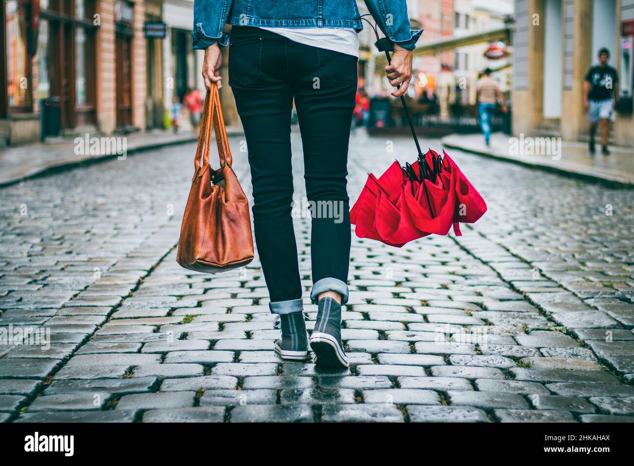 Donna che tiene la borsa di pelle e ombrello rosso e sta camminando sulla strada dopo la pioggia. Concetto di moda in città. Donna alla moda che indossa abiti casual. Foto Stock