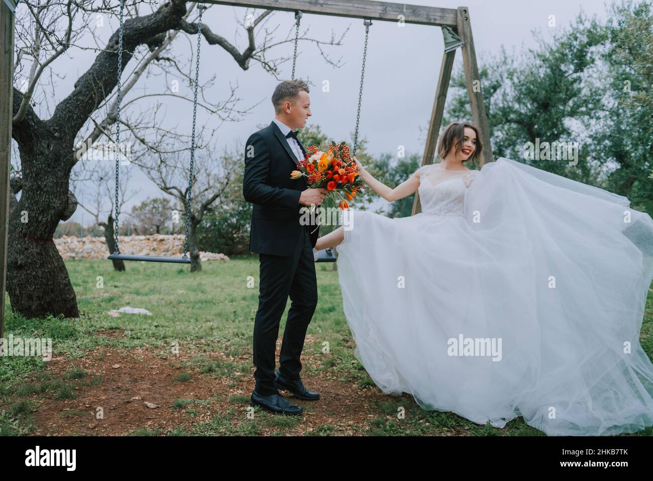 Felice coppia sorridente di stile che cammina in Toscana, Italia il giorno del matrimonio. SPOSI NOVELLI CON L'ALTALENA NEL PARCO. La sposa e lo sposo camminano giù il Foto Stock