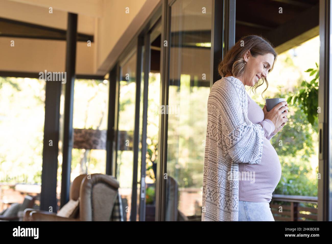 Donna incinta caucasica che tiene una tazza di caffè sorridente mentre guarda fuori dalla finestra a casa Foto Stock