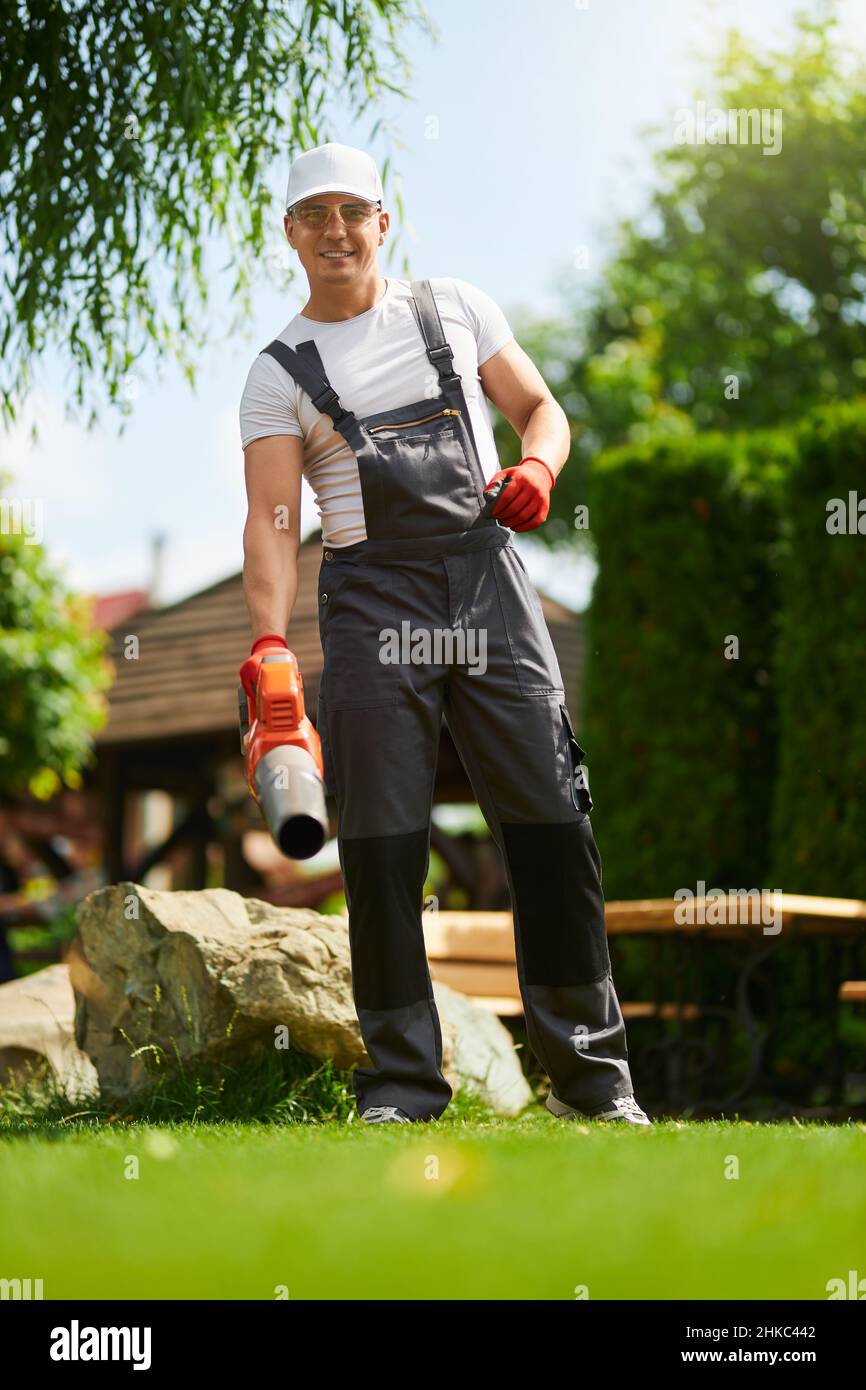 Giardiniere maschile muscoloso in uniforme, cappello e guanti in piedi sul prato verde, sorridendo e guardando la macchina fotografica mentre si utilizza il soffiatore foglie all'aperto. Processo di lavoro durante l'estate. Foto Stock