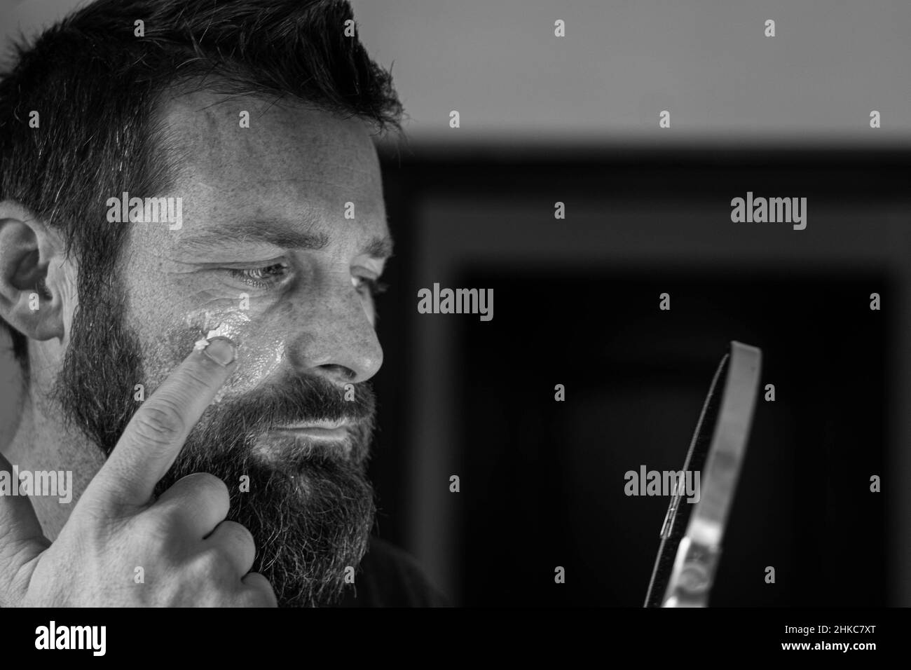 Un giovane uomo sta applicando dopo aver pulito il suo viso giorno o notte crema per prendersi cura della pelle dietro uno specchio in un bagno. Foto Stock