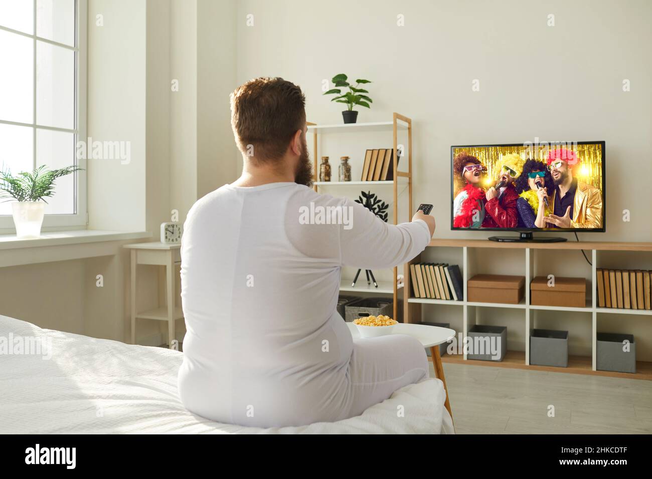 Ragazzo divertente chubby in pigiama seduto in camera da letto e guardare la televisione in televisione Foto Stock