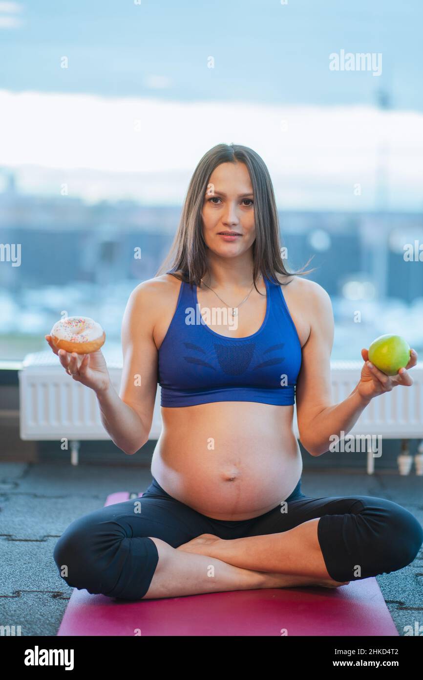 Giovane donna incinta beauteous in abbigliamento sportivo blu si siede su tappeto rosa scegliendo tra mela e ciambella in palestra. Verticale. Foto Stock
