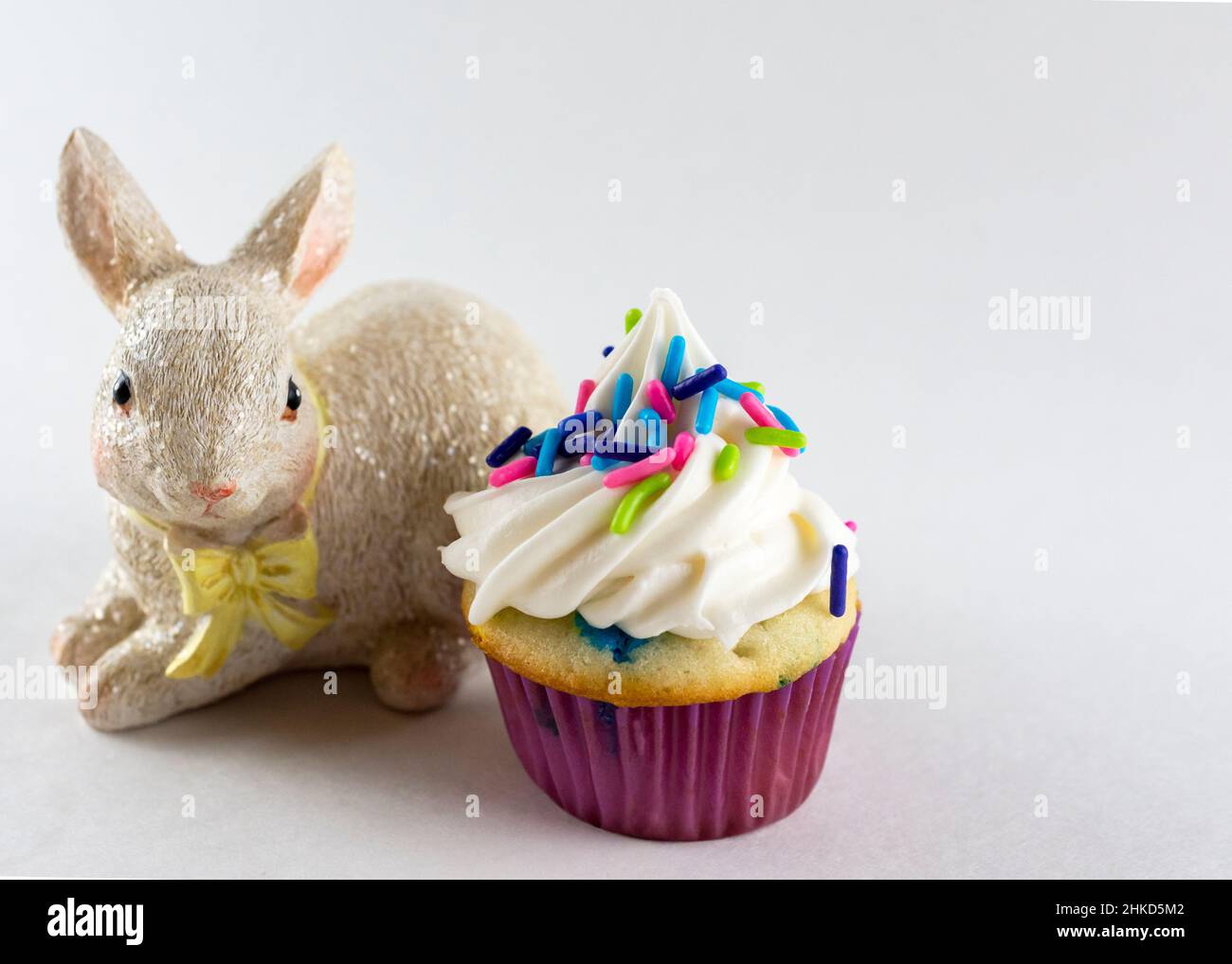 Vista ravvicinata di un mini cupcake decorato con mini spolverini di caramelle color primavera. Bunny figuringe in background. Sfondo bianco con copia s. Foto Stock