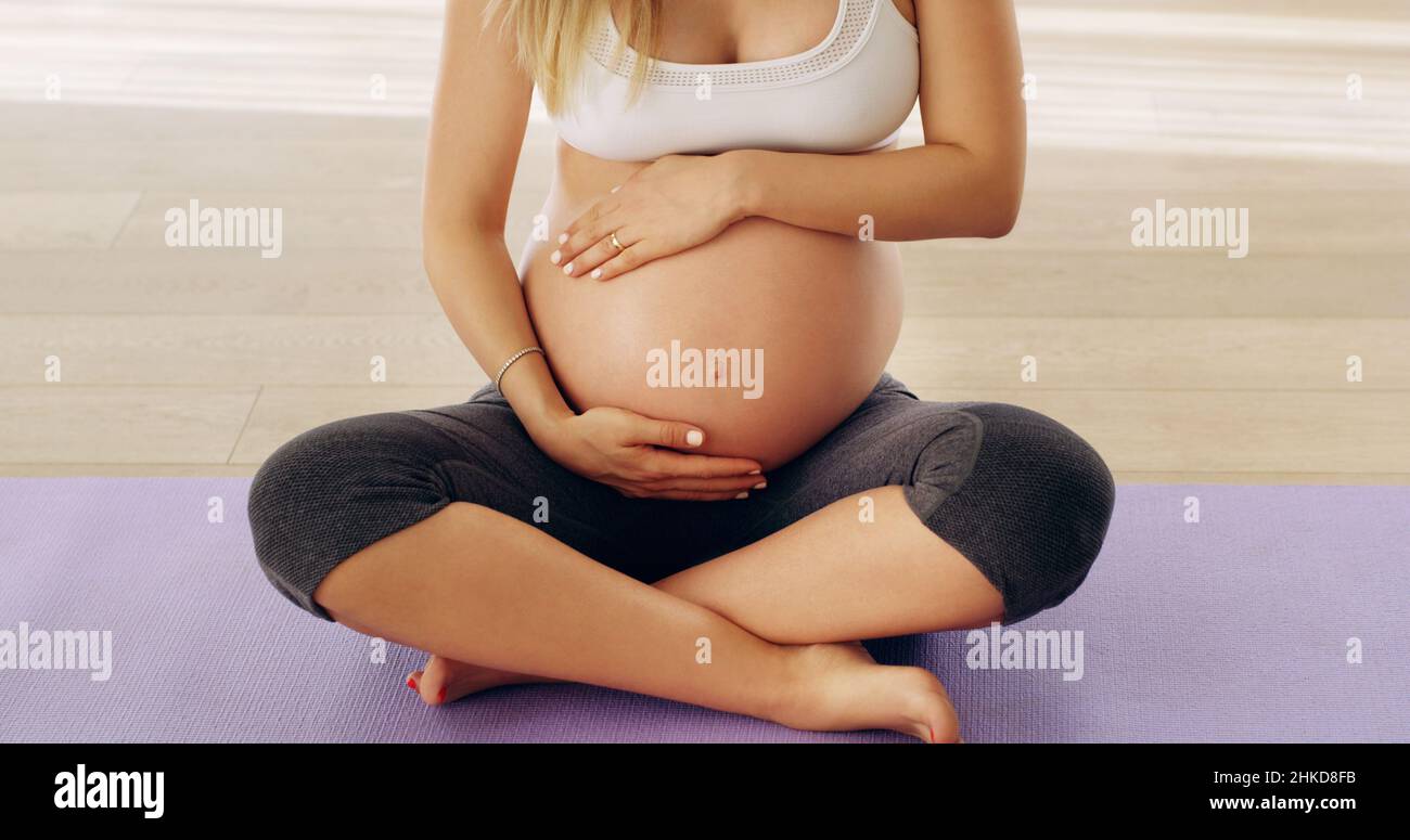 Non prendetevi cura di voi. Scatto corto di una giovane donna incinta irriconoscibile strofinando il suo bump del bambino mentre si siede su un tappeto di yoga. Foto Stock