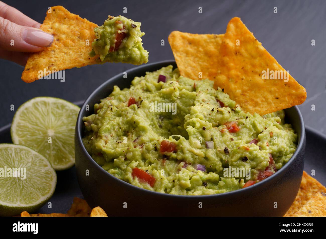 Primo piano di mano donna con tortilla chips o nachos con gustoso guacamole fresco tuffo in piatto nero su sfondo nero Foto Stock