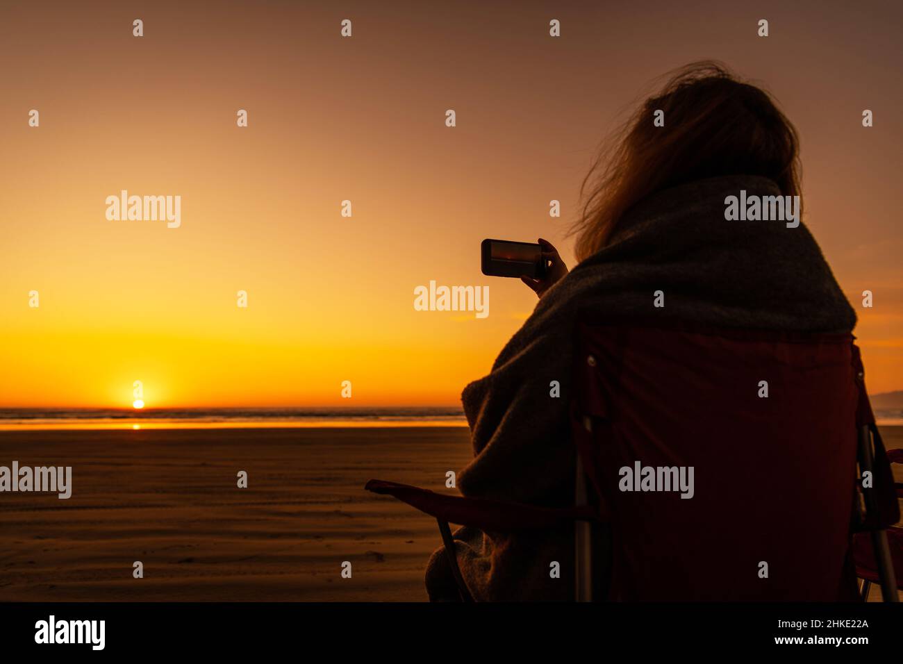 Donna caucasica coperta da una calda coperta che prende il video di un tramonto Scenic Beach mentre si siana su una sedia del campo. Tema Vacanze fronte Oceano. Foto Stock