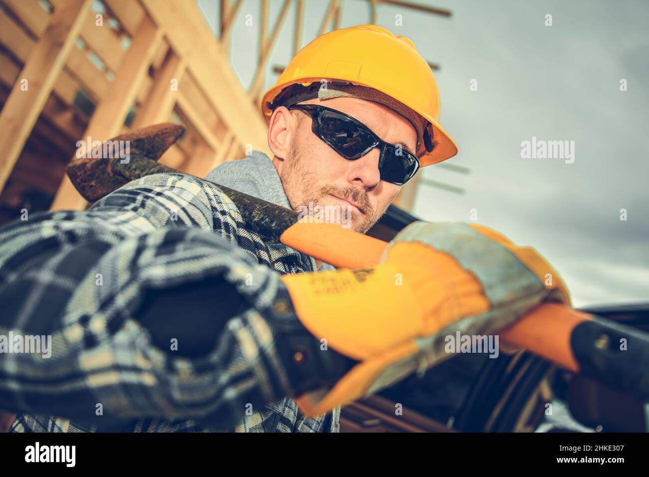 Caucasico professionista Construction Contractor Worker nel suo 40s con un martello grande sulla spalla. Indossare cappello rigido giallo e protezione occhi Sungla Foto Stock