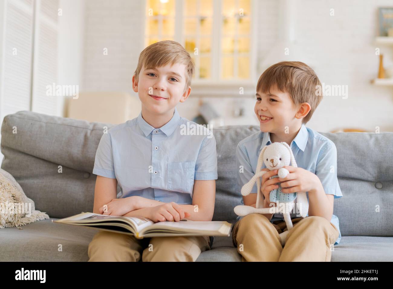 Due ragazzi seduti sul divano stanno leggendo un libro. Il fratello maggiore legge storie interessanti al fratello minore, concetto di amicizia, intelligenza, interazione, fratello minore tiene il coniglio giocattolo in mano Foto Stock