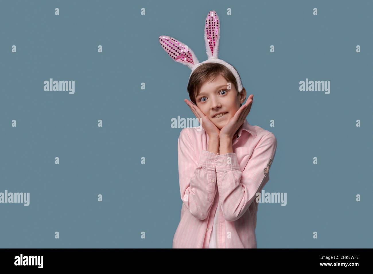 Un giovane ragazzo in camicia rosa con orecchie di coniglietto pasquale sulla testa si pone allegro e gesticula con le mani in studio su uno sfondo blu. Felicità e gioia dalle vacanze primaverili Foto Stock