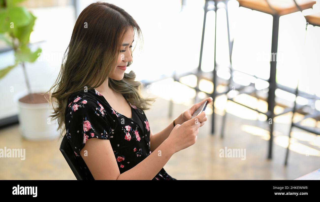 Bella donna millenaria asiatica che usa il suo smartphone in un caffè luminoso minimalista. Guarda video online e gioca ai videogame mobili. Foto Stock
