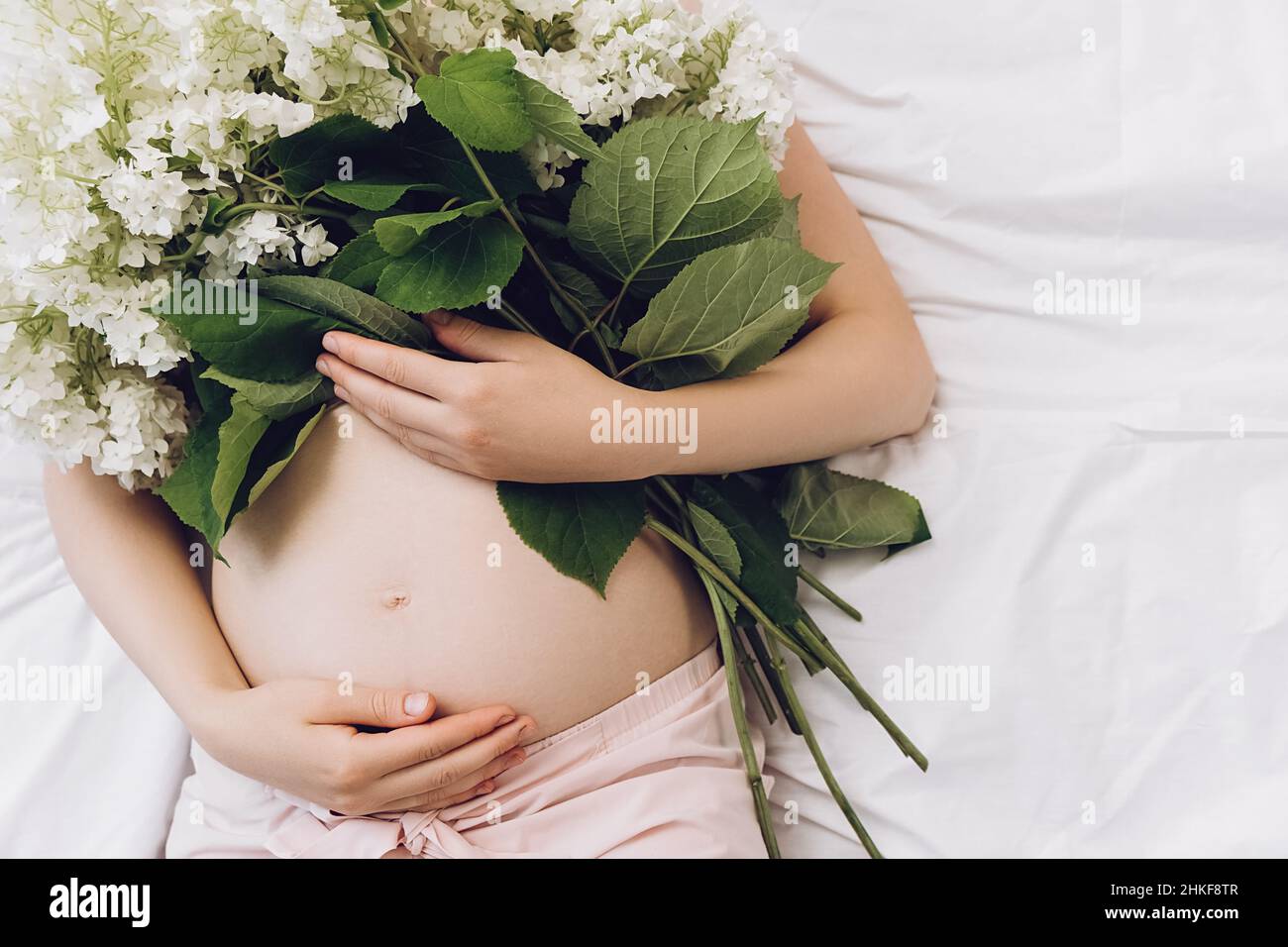 Primo piano di giovani donne incinte mani toccare delicatamente pancia sdraiato sul comodo letto che tiene bei fiori freschi estate. L'aspettativa della futura madre Foto Stock