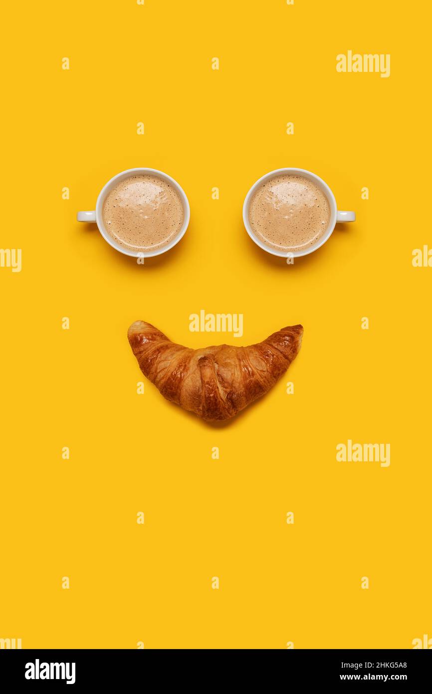 Due tazze di caffè con schiuma e un croissant che formano un viso sorridente su sfondo giallo con spazio per la copia e spazio per il testo Foto Stock