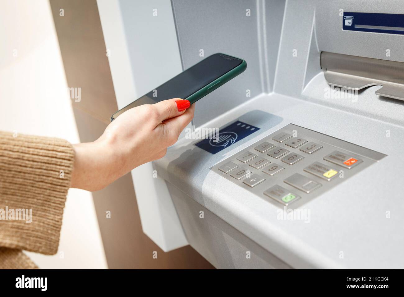 Mano femminile con telefono cellulare che preleva denaro da atm utilizzando il sistema a pagamento Wi-fi senza contatto NFC. Autenticazione wireless e trasmissione dati Foto Stock