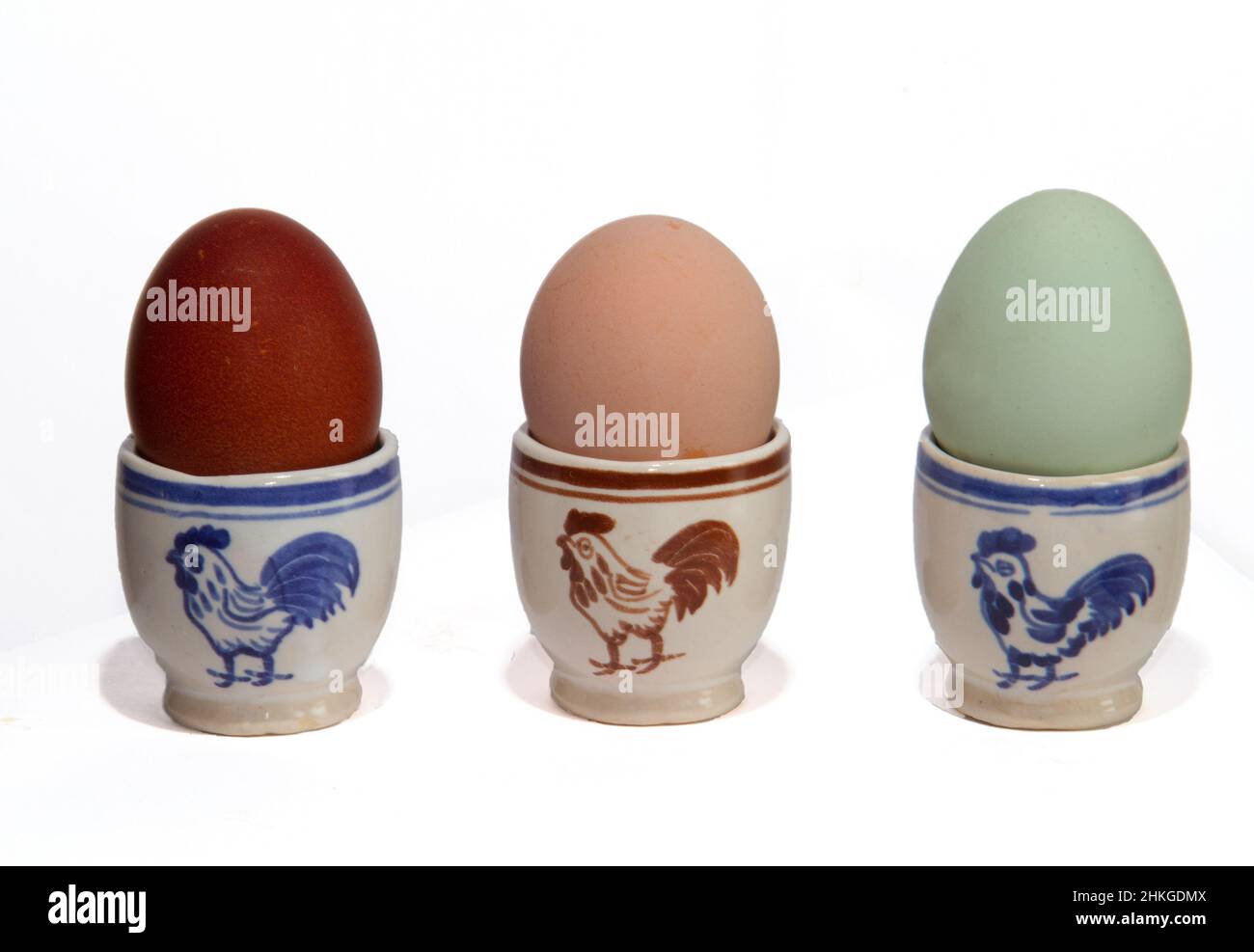 Tre coppe d'uovo, decorate con un gallo, uova in tre colori diversi, posate da tre tipi di polli: Sussex, Marans e Araucona Foto Stock