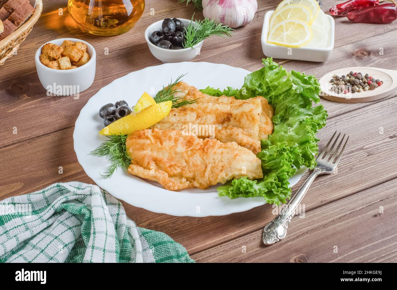 Pezzi di filetto di pesce fritto con limone ed erbe su un piatto bianco su sfondo di legno scuro. Foto Stock